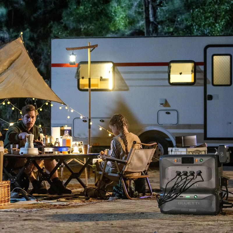 Two people sitting at a table outside an RV with string lights and a Bluetti Apex 300 power station.